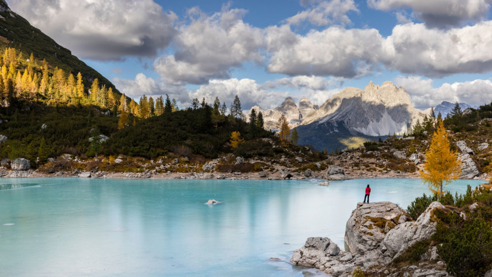Lago di Sorapis en automne. Les arbres sont jaunes et le lac est gelé. Un personnage est debout sur un rocher.