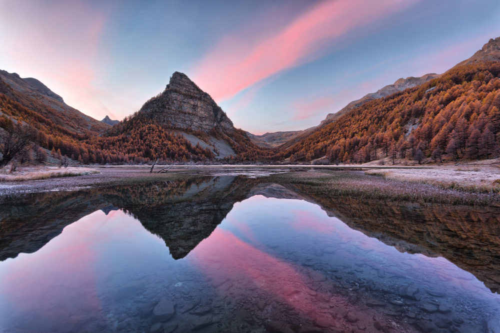 Le Lac des Sagnes en automne au petit matin. Les arbres sont jaunes et les nuages roses.